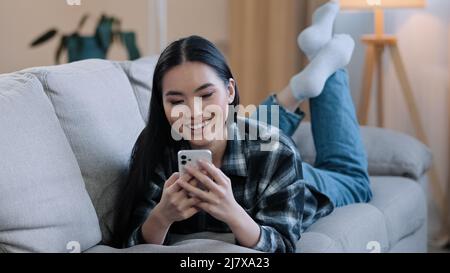 Asiatische weibliche entspannende Mädchen Frau auf der Couch zu Hause liegen genießen Chat Eingabe App Handy entspannen beobachten virale live Video spielen Spiel folgen wie post in Stockfoto