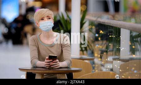 Kaukasische Erwachsene Frau blond in medizinische Maske reife weibliche Besucher sitzt im Café am Tisch schaut auf Telefon Checks Zeit auf dem Smartphone Blick zur Seite Stockfoto