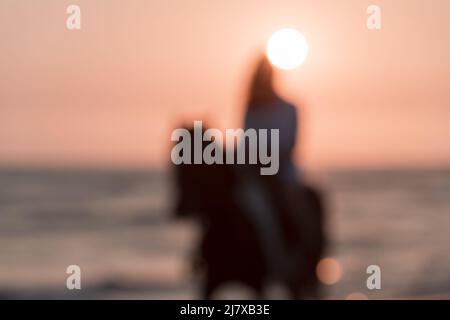 Verschwommenes Foto. Frau in Sommerkleidung genießt es, bei Sonnenuntergang auf einem Pferd an einem schönen Sandstrand zu reiten. Selektiver Fokus Stockfoto