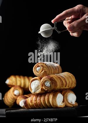 Gebackene knusprige Röhrchen mit Proteinfüllung, bestreut mit Puderzucker auf einem schwarzen Tisch, Dessert Stockfoto