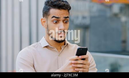 Porträt bärtig jung erstaunt stattlichen Geschäftsmann steht in der Stadt suchen im Handy mit schockiertem Ausdruck auf Gesicht liest schlechte Nachrichten gut Stockfoto