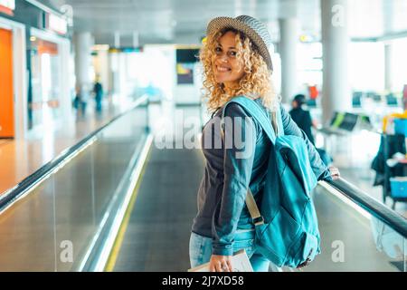 Fröhliche junge Erwachsene Frau reist am Flughafen mit Rucksack. Weibliche Menschen porträtieren lächelnd und gehen am Tor. Ziemlich blonde Dame Reisende en Stockfoto