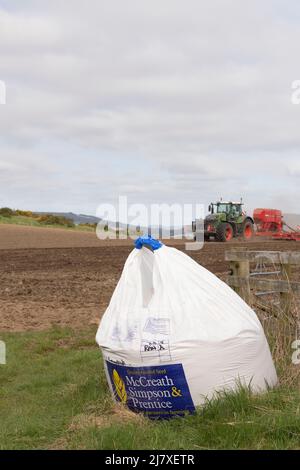 Ein Beutel Gerstensamen an einem Feldtor im Frühjahr, mit einem grünen Traktor, der einen Sämaschinen-Bohrer schleppt, der im Hintergrund in Betrieb ist Stockfoto