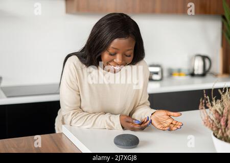 Afroamerikanische 30s-Mann-Frau, die zu Hause mit dem digitalen virtuellen Assistenten spricht, eine Frage stellt oder um einen Musikwechsel bittet. Intelligentes AI-Lautsprecherkonzept und Sprachsteuerung Stockfoto