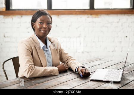 Intelligente Büroangestellte, die vor die Kamera schaut, am Schreibtisch vor dem Laptop sitzt, eine erfolgreiche afroamerikanische Geschäftsfrau, die gerne ihren Job liebt und bereit für die nächste Herausforderung ist Stockfoto