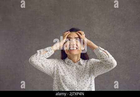 Unglückliche, verrückte Frau, die auf einem grauen Hintergrund steht, schreit und weint, während sie ihren Kopf hält. Stockfoto