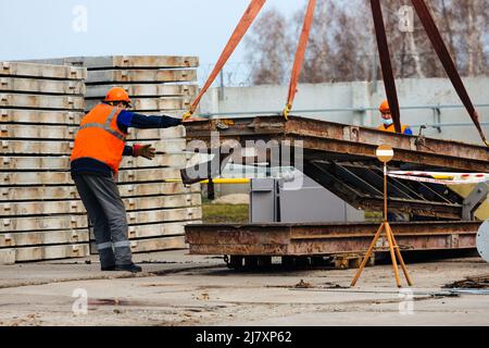 Slinger in Helm und Weste steuert das Entladen von Metallkonstruktionen auf der Baustelle. Weißer Handwerker entlädt Ladung. Stockfoto