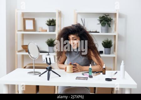 Fröhlich gebräunt schöner lockiger lateinischer Beauty-Blogger in Casual Top erzählt von Körpercreme im Heim-Interieur. Space Mockup Banner kopieren. Influencer-Rekord Stockfoto