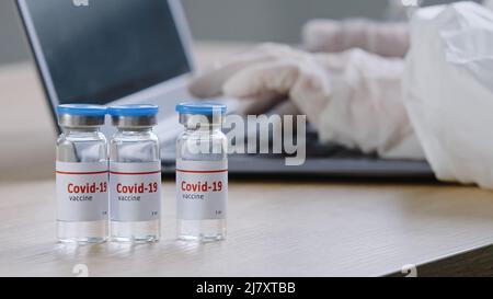 Nahaufnahme von covid-19-Impfstoff steht auf Holztisch in der Klinik unkenntlich Arzt Arzt Arzt Arbeiter Wissenschaftler in Latexhandschuhe tippen auf Laptop macht Stockfoto