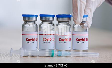 Nahaufnahme Detail von vier Glasflaschen mit covid19 Impfstoff gegen Coronavirus Pandemie stehen auf Holztisch in einem Labor in der Nähe von Spritze Hand in Stockfoto