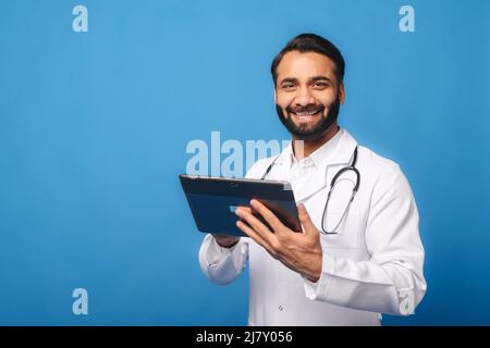 Lächelnder indischer Arzt, Pädiatrie, Physiotherapeut, trägt ein weißes Kleid mit Stethoskop auf den Schultern und nutzt ein digitales Tablet für die Online-Kommunikation mit Patienten, die auf blauer Telemedizin isoliert sind Stockfoto