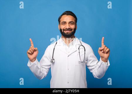 Indischer Arzt, Pädiatrie, Arzt, Therapeut trägt einen weißen medizinischen Kittel mit einem Stethoskop auf den Schultern zeigt die Finger nach oben auf leeren Raum, Mock-up, Copy Space. Gesundheitswesen und Medizin Stockfoto