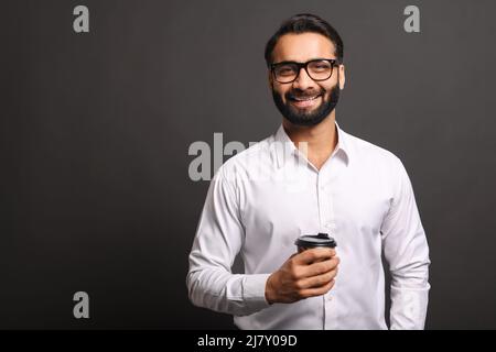 Ein gutaussehender, selbstbewusster bärtiger Inder in formeller Kleidung steht mit einer Tasse Kaffee zum Mitnehmen isoliert auf Schwarz, ein lächelnder, männlicher Büroangestellter in einer Brille nimmt eine Kaffeepause ein Stockfoto