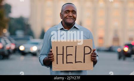 Unglücklicher schwarzer afroamerikanischer Geschäftsmann, der Karton mit Text hält, hilft beim emotionalen Zeigen auf dem Schild. Gestresst frustriert Mann halten Poster suchen Stockfoto