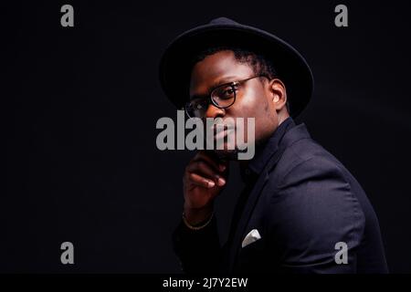 Junger hübscher afro-amerikanischer melandiöser Junge in einer Jacke und einer Brille auf schwarzem Hintergrund im Studio.Mysterium und Trauer verstecken sich Stockfoto