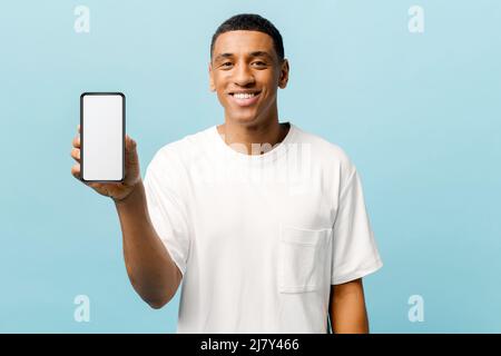 Glücklicher afroamerikanischer junger Mann, der auf blauem Hintergrund ein Telefon mit leerem Bildschirm zeigt, für neue mobile App, ein Mockup-Konzept, ein Smartphone mit leerem Display in den Händen eines jungen Mannes Stockfoto