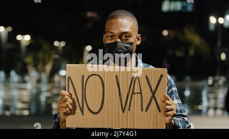 Junger wütender afroamerikanischer Mann in schwarzer Maske steht nachts in der Stadt und hält ein Schild mit dem Text No VAX guy Protest gegen den Impfstoff Covid 19 Stockfoto