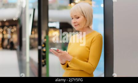 Lächelnde ältere kaukasische Frau im mittleren Alter, die das Telefon hält und auf den Smartphone-Bildschirm schaut, um eine Nachricht zu senden, die mithilfe von mobilen Anwendungen E-Mails absieht Stockfoto