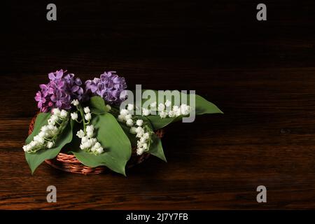 Schönes Bouquet von Maiglöckchen und lila Flieder in braunem Weidenkorb auf schwarzem Hintergrund. Visitenkarte. Stockfoto