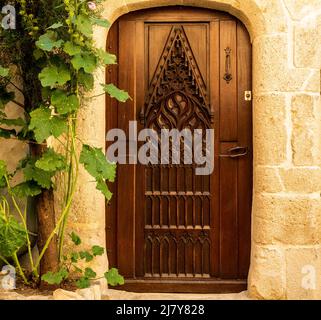 Eingangstür aus kunstvoll und reich eingelegtem dunklem Holz, von der Straße in der Provence, Frankreich, eingerahmt von beigefarbener Steinmauer und einem grünen Zweig Stockfoto