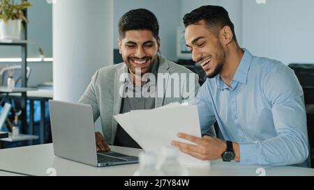 Lächelnde junge Kollegen mit Papierdokumenten, indische Mitarbeiter sitzen mit einem Laptop im Büro und erhalten gute Nachrichten. Zwei erfolgreiche arabische Männer Stockfoto