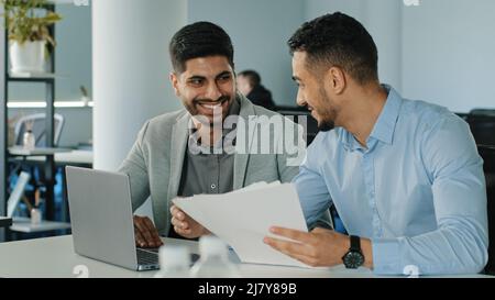 Zwei erfolgreiche arabische männliche Geschäftsleute, die im Büro sitzen und Papierdokumente halten. Bunt lächelnde junge Kollegen, indische Mitarbeiter, die sich einen Laptop angeschaut haben Stockfoto