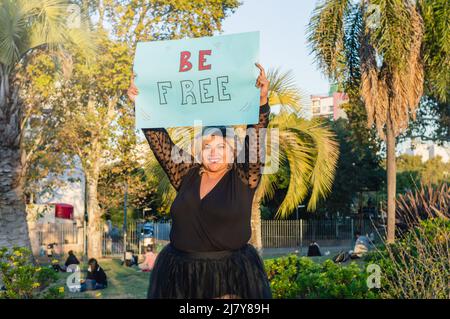 Plus size kaukasische Frau, in schwarz gekleidet, im Park mit einem Schild mit der Aufschrift „frei sein“, lächelnd und schauend auf die Kamera. Stockfoto