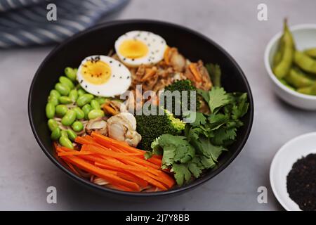 Draufsicht auf Schale mit japanischen udon-Nudeln, würzigen Jakobsmuscheln, Eiern, Karotten, Bohnen und Sesamsamen auf schwarzer Schale und grauem Betonhintergrund. Flach liegend Stockfoto