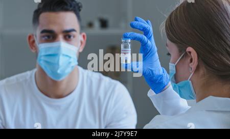 Blick von hinten kaukasische unkenntlich Frau Arzt trägt medizinische Handschuhe hält Glas mit covid-19 Virus-Impfstoff-Medikament zeigt arabischen spanier Kerl Mann Stockfoto