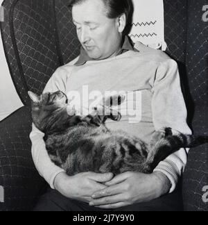 1960s, historisch, ein Mann, der auf einem Liegestuhl sitzt und seine Katze in den Armen liegt und auf sein Gesicht blickt, England, Großbritannien. Stockfoto