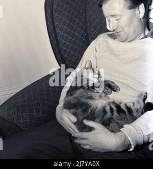 1960s, historisch, ein Mann, der in einem Liegestuhl sitzt und seine Katze auf dem Schoß hält, England, Großbritannien. Stockfoto