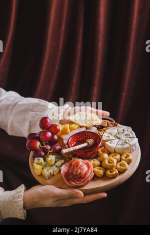 Eine Frau hält in ihren Händen ein rundes Holzschneidebrett mit Wein-Vorspeisen - Schneiden von Käse, Wurst und Obst auf dem Hintergrund aus Stoff Stockfoto