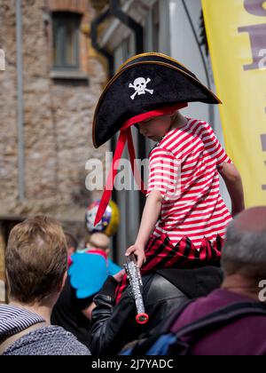 Kleiner Junge in Piratenkostüm, der auf den Schultern des Vaters beim Brixham Pirate Festival 2022 in Devon, Großbritannien, getragen wird Stockfoto