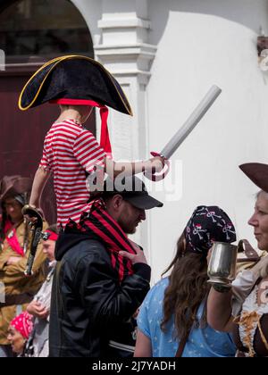 Kleiner Junge in Piratenkostüm, der auf den Schultern des Vaters beim Brixham Pirate Festival 2022 in Devon, Großbritannien, getragen wird Stockfoto