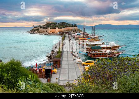 Alte Burg auf Pigeon Island (Guvercinada) in Kusadasi Türkei bei Sonnenuntergang mit bunten Booten entlang des Hafens. Stockfoto