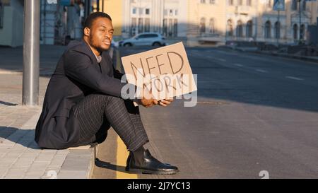Verzweifelte verärgerte afroamerikanische Millennial Student trägt Anzug sitzen auf Treppen im Freien halten Plakat brauchen Arbeit, schwarzer Geschäftsmann nervös Stockfoto