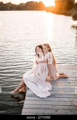 Eine Schwangerin mit ihrer Tochter vor dem Hintergrund des Wassers bei Sonnenuntergang, eine Frau mit einem Bauch genießt den Sonnenuntergang am Fluss Stockfoto