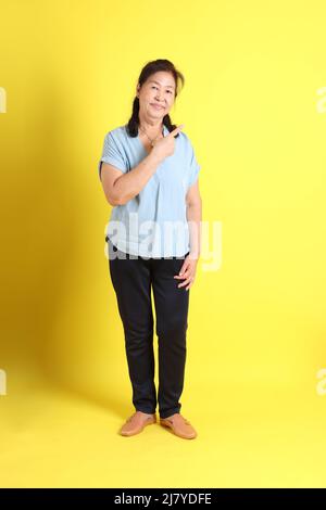 Die asiatische ältere Frau trägt ein hellblaues Hemd, das auf dem gelben Hintergrund steht. Stockfoto