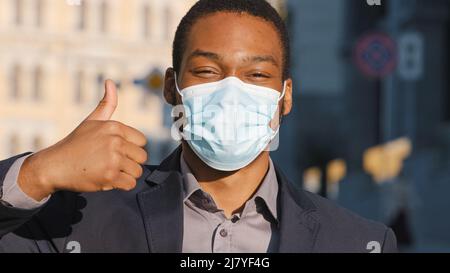 Gemischter Geschäftsmann in Anzug trägt medizinische Gesichtsmaske. Nahaufnahme der Hand, die den Daumen nach oben zeigt. Vorbeugende Vorsichtsmassnahmen aufgrund von Covid-19 Stockfoto