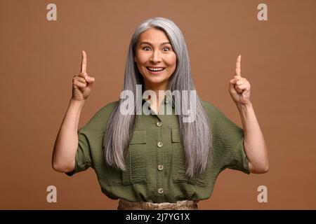 Lächelnde ältere grauhaarige Geschäftsfrau, isoliert auf grauem Hintergrund, zeigt den Finger nach oben. Dame, die auf etwas achtet Stockfoto