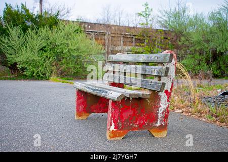 Abgenutzte rote und graue Holzbank am Rand einer Schotterstraße mit einem verlassenen Gebäude im Hintergrund Stockfoto