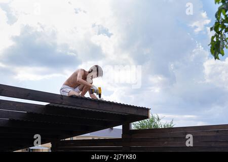 Gebäudedach. Junge Männer reparieren das Dach. Dachkonstruktion. Arbeiten in der Höhe mit einem Schraubendreher. Stockfoto