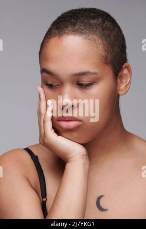 Traurige, schöne junge afroamerikanische Frau mit kurzen Haaren, die an Krebs erkrankt ist und sich mit dem Kopf an der Hand lehnt Stockfoto