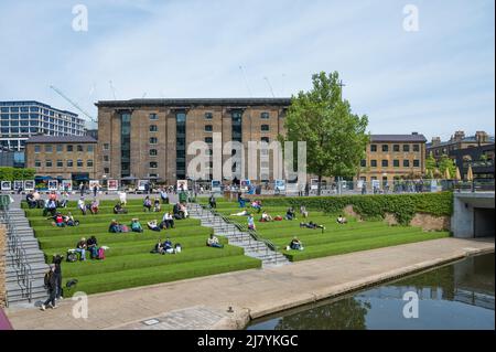 An einem sonnigen Frühlingstag im Coal Drops Yard entspannen sich die Menschen auf der Rasenterrasse am Ufer des Regent-Kanals. London, England, Großbritannien. Stockfoto