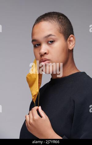 Porträt einer kurzhaarigen jungen schwarzen Frau mit gelber Maske, die auf dem Ohr vor grauem Hintergrund hängt Stockfoto