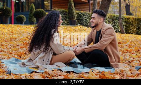 Fröhliches hispanische Paar auf Datum im Herbst Park junge Hände halten lachend und lächelnd sitzen gekreuzt auf karierten in bunten Blättern im Freien Stockfoto