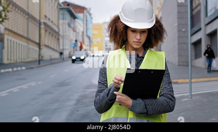 Portrait junge afroamerikanische schöne Ingenieurin mit Sicherheitsweste und Hardhut in der Stadt in der Nähe des Straßenverkehrs, die Daten schreibt Stockfoto
