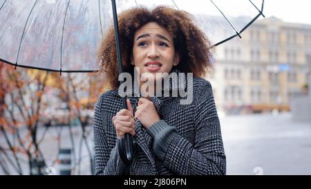 Afroamerikanisches Mädchen lockig-haarige traurige Frau in stilvollem Mantel steht im Herbst auf der Stadtstraße mit transparentem Regenschirm im Regen leidet unter kalten Winden Stockfoto