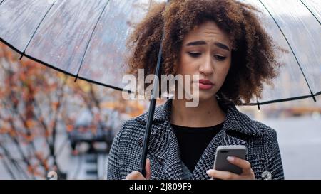 Afroamerikanische Frau lockiges Mädchen mit transparentem Regenschirm steht auf Herbststraße im Regen schaut auf Handy aufgeregt Traurigkeit besorgt über niedrig Stockfoto