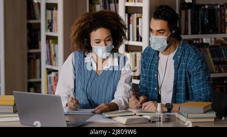 Zwei Studenten in medizinischen Masken sitzen am Schreibtisch in der Universitätsbibliothek hören Lehrer in Kopfhörer auf Laptop Studie notieren Informationen in Notebook Stockfoto